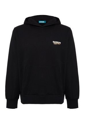 Joven con cabello rizado y castaño, vistiendo una sudadera negra con el logo "Barrow" en el pecho y pantalones beige, de pie frente a un fondo blanco.