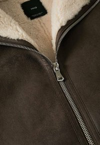 Giacca in pelle marrone con cerniera argentata e foderatura in pile color crema. Etichetta visibile sul colletto. Superficie texturizzata.