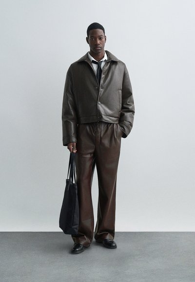 Moški stoji ob navadni steni, oblečen v temno rjavo usnjeno jakno in hlače, belo srajco, črno kravato, črne čevlje, ter drži črno torbo.