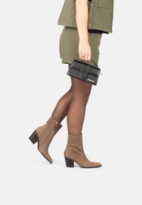 Botas de ante marrón hasta el tobillo con punta afinada y tacón cuadrado, combinadas con un chaleco acolchado verde y un bolso negro.