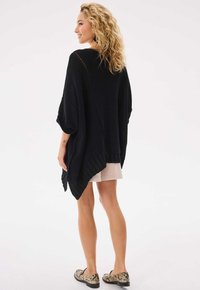 Czarny dziany ponczo z szerokimi rękawami, z teksturowanym wzorem i asymetrycznym dołem. W zestawie z jasną spódnicą i butami w wężowy wzór.