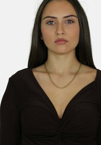 Top nero con scollatura profonda, caratterizzato da un dettaglio annodato al centro. Una collana spessa in oro riposa sulla clavicola.