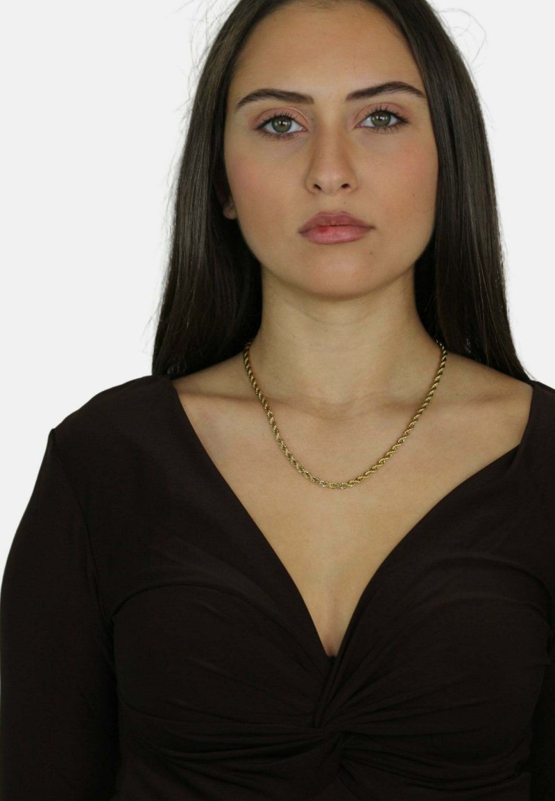 Top nero con scollatura profonda, caratterizzato da un dettaglio annodato al centro. Una collana spessa in oro riposa sulla clavicola.