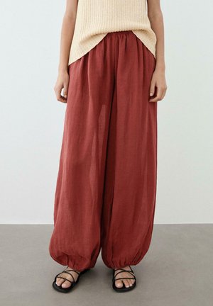 Frau trägt locker sitzende, rostfarbene Hose mit elastischen Bündchen, beiges ärmelloses Stricktop und schwarze Riemchensandalen, steht auf grauem Boden.