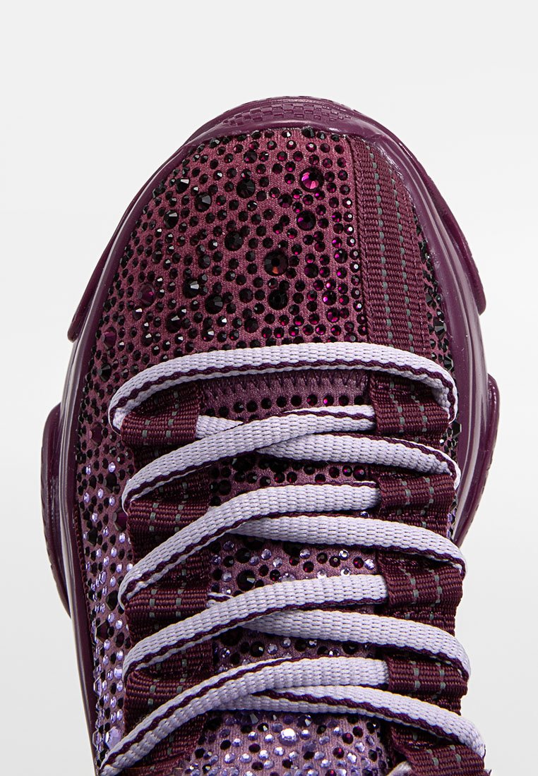 Zapatilla morada con un upper texturizado y acentos brillantes, cordones blancos y una suela de goma redondeada. El diseño multicolor mejora la visibilidad.