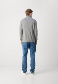 Pull en tricot gris avec des poignets et un ourlet côtelés, porté avec un jean en denim bleu clair et des chaussures de couleur crème. Fond uni.