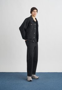 Casaco de ganga preto, calças pretas combinando e ténis cinzentos. Modelo está de pé numa superfície azul com um fundo branco liso.