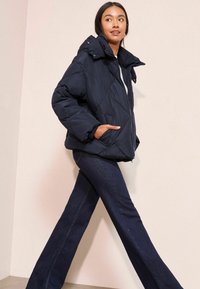 Nafukovací bunda v námořnické modři se zapínáním na zip, oversized límcem a prošívaným vzorem; v kombinaci s rozšířenými tmavými džíny.