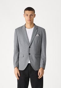 Grå blazer med en strukturerad mönster, en bröstficka med en mönstrad näsduk, tvåknapplås, och slagkrage, som matchas med en vit t-shirt.