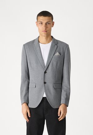 Blazer gris avec un motif texturé, poche poitrine unique avec un mouchoir à motifs, fermeture à deux boutons et revers crantés, associé à un t-shirt blanc.