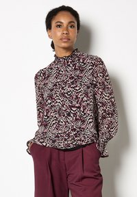 Blusa a maniche lunghe con un alto colletto arricciato, caratterizzata da un motivo astratto viola e beige. Realizzata in tessuto leggero, leggermente arricciata ai polsini.