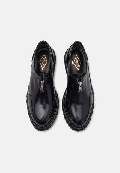 Black glossy leather slip-on shoes with a zippered front and rubber soles featuring ridged edges. Gold logo on the insole.