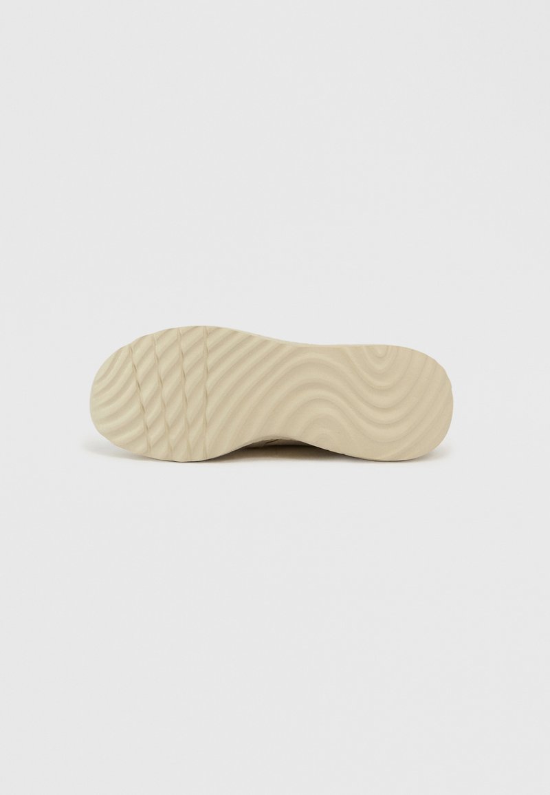 Suela de zapato de goma en beige claro con una textura ondulada y ranuras verticales para mayor tracción. Perfil plano y diseño de puntera redondeada.