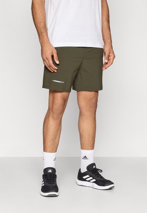 Olijfgroene sportshorts met een ontspannen pasvorm, voorzien van een reflecterende streep, gecombineerd met zwarte sportschoenen en witte sokken.