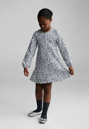 Blumendruck-Kleid mit langen, gepufften Ärmeln, gerafftem Oberteil und ausgestellt Saum. Die Farben umfassen Weiß, Marineblau und Hellgrau. Kombiniert mit grauen Socken und silbernen Schuhen.