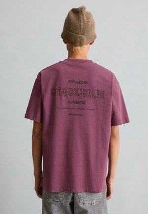 Person trägt ein lila T-Shirt mit "Founded in Stockholm Authentic Scandinavian denim brand" auf dem Rücken und eine braune gestrickte Mütze.