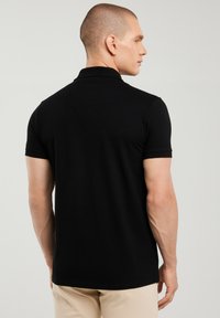 Mann mit kurzen Haaren, der ein schwarzes kurzärmeliges Polohemd und beige Hosen trägt, von hinten gesehen mit dem Kopf nach rechts gedreht.