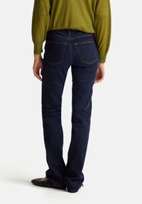 Jeans acampanados de color azul oscuro con bolsillos traseros distintivos y costuras en contraste, combinados con una blusa verde y zapatos planos negros.