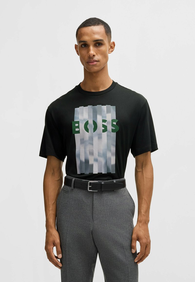 Schwarzes T-Shirt mit einem großen Grafiken-Druck in Grau und Grün, auf dem das Wort "BOSS" zu sehen ist. Das Model trägt graue Hosen und einen schwarzen Gürtel.