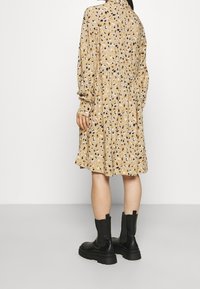 Robe beige avec un motif tamisé multicolore et ludique ; présente des manches longues, un col et une taille froncée ; associée à des bottines noires à la cheville.