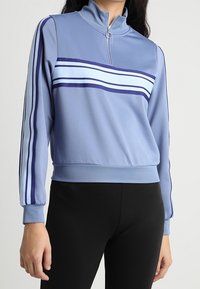 Giacca zip-up azzurro chiaro con colletto alto, righe orizzontali blu scuro e blu chiaro, e maniche arricciate. Texture del tessuto liscia.