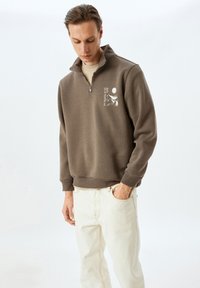 Brauner Reißverschluss-Sweatshirt mit erhöhtem Kragen, das ein grafisches Design auf der linken Brust zeigt. Kombiniert mit hellcremefarbenen Hosen. Glatte Textur.