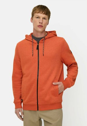 Ung mand iført en orange hættetrøje med lynlås og snøre samt beige bukser, stående med den ene hånd i lommen.