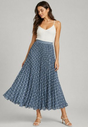 Femme portant un haut blanc sans manches et une jupe midi plissée bleue à pois blancs, debout devant un fond uni.