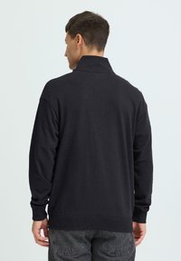 Pull à manches longues sombre avec un col montant, fabriqué en tissu doux. Présente des poignets et un ourlet côtelés, ainsi qu'une coupe décontractée.