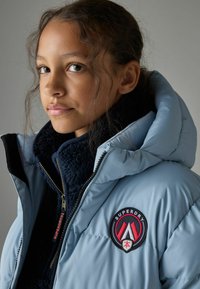 Veste rembourrée bleu clair avec une doublure intérieure en fourrure navy, dotée d'un patch logo et d'une grande capuche, fermeture éclair et détails cousus.
