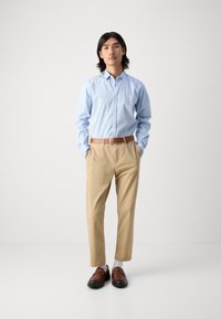 Chemise bouton-bleue claire, chinos beiges, ceinture marron et mocassins marron foncé. Le mannequin se tient droit avec les mains dans les poches. Design simple, sans motifs.