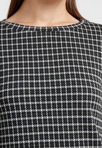 Černobílá kostkovaná látka s houndstooth vzorem, s kulatým výstřihem a měkkou texturou. Detailní pohled zdůrazňuje design.