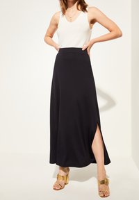 Svart maxikjol i mjukt tyg med en sidslits, kombinerad med en figurnära vit linne och gyllene klacksandaler. Slät textur, enkel design.