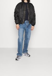 Svart bomberjacka med satinfinish, ribbade cuffs, guldfärgad dragkedja, ihopparad med blå jeans och lätta grå sneakers.