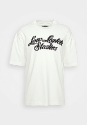 Hvid t-shirt med "Low Light Studios" trykt i sort skrift, kombineret med beige bukser. Placeret ved siden af en kopimaskine med grå og hvide detaljer.