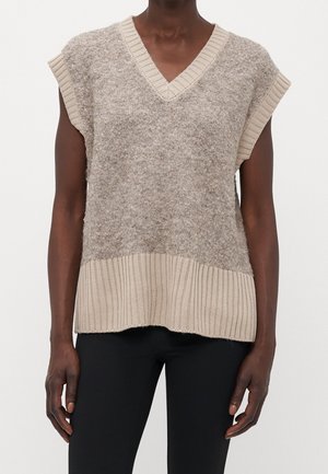 Personne à la peau foncée portant un gilet sans manches beige en tricot avec un col en V et un pantalon noir sur un fond blanc uni.