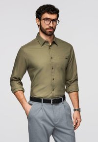 Homme avec barbe et lunettes portant une chemise kaki à boutons aux manches retroussées et un pantalon gris, posant avec une main dans la poche.