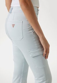 Pantaloni cargo azzurro chiaro con tasche posteriori, caratterizzati da un piccolo logo a triangolo rosso. Il materiale è liscio e elastico, con dettagli di cucitura visibili.