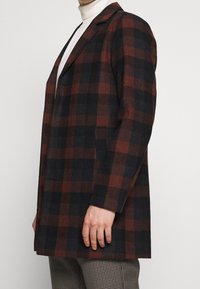 Kostkovaný kabát v tmavě hnědé a černé barvě se strukturovaným designem, s jedním řadovým zapínáním a dvěma postranními kapsami. Měkký materiál ze směsi vlny.