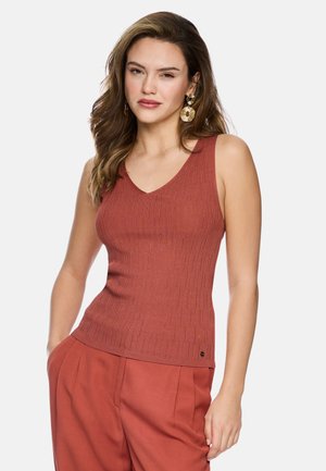 Junge Frau mit lockerem welligem Haar, die ein rostfarbenes, geripptes, ärmelloses Oberteil und passende Hose trägt, ergänzt durch große runde Ohrringe.