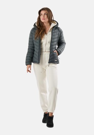 Femme portant une doudoune gris foncé par-dessus un sweat à capuche zippé beige clair et un pantalon de jogging assorti, debout devant un fond blanc.