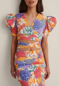 Robe fleurie avec un décolleté en V profond, des manches bouffantes, des teintes vives de violet, orange et vert, dotée de découpes et de détails froncés.