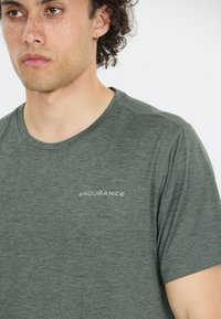 Camiseta de manga corta verde oscuro hecha de tejido transpirable. Presenta un escote redondo y el logo "ENDURANCE" en gris claro en el pecho.