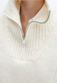 Nahaufnahme eines weißen gestrickten Pullovers mit silbernem Reißverschluss, der teilweise geöffnet ist, sodass ein weißes Hemd mit Kragen darunter sichtbar wird.