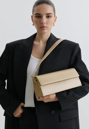 Femeie îmbrăcată într-un sacou negru și un top alb, care ține o geantă bej dreptunghiulară de umăr cu clapetă, pe un fundal simplu.