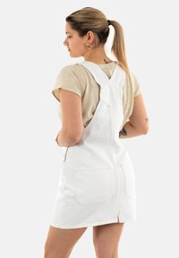 Vestido de mezclilla blanca tipo mono con forma ajustada, dos grandes bolsillos traseros y tirantes ajustables. La tela parece texturizada y resistente.