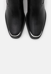 Botines de cuero negro hasta el tobillo con punta afilada, que presentan un elegante borde plateado en la base y detalles de costura en los laterales.