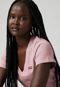 Jasnoróżowa koszulka typu V-neck z krótkimi rękawami, z czerwonym naszywką z logo Levi's po lewej stronie. Gładki materiał i luźny krój.