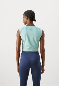 Svjetlo tirkizni bezrukavni crop top s teksturiranim prednjim dijelom i logom "NIKE PRO". Uparen s mornarsko plavim tajicama visokog struka. Glatka, sportska tkanina.
