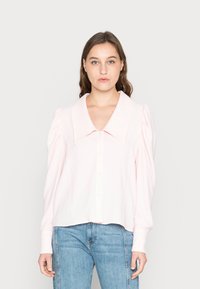 Camisa de botão rosa claro com um grande colarinho, mangas bufantes e tecido texturizado. Combinada com jeans azul claro.